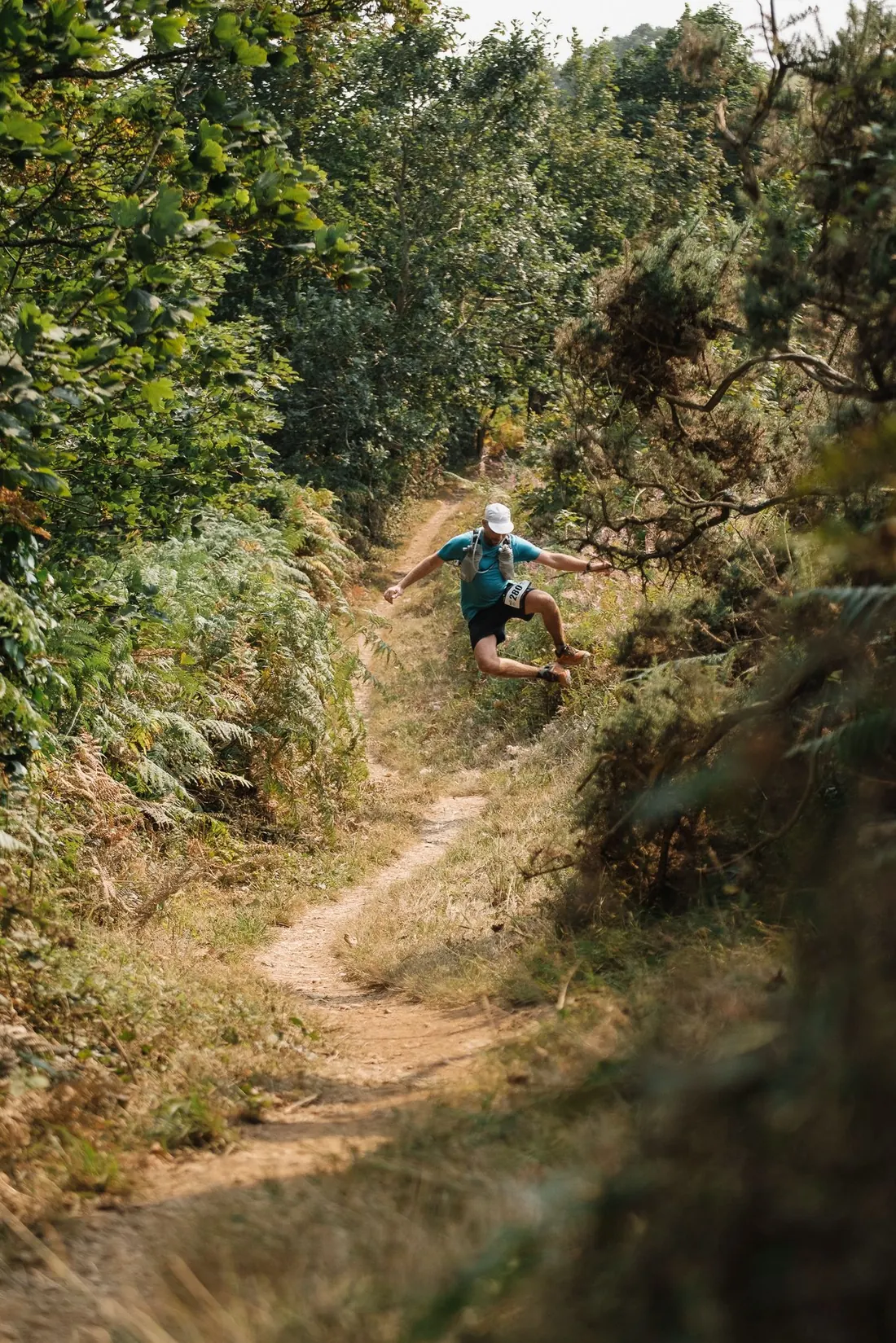 A PZ×RC runner mid-air over a rut in a sunken coast-path track, arms out, bracken either side