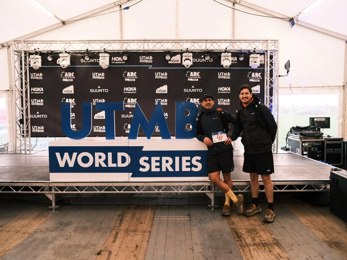 Two runners in front of the UTMB World Series backdrop in the Arc of Attrition event marquee, one still wearing an Arc 100 race number