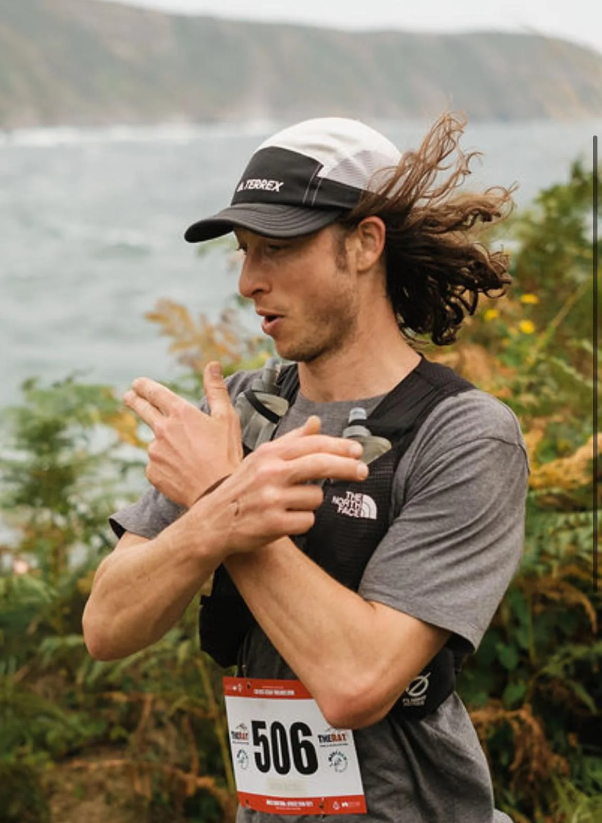 A runner adjusting a sleeve on an exposed cliff path, hair blown sideways, sea and headland behind