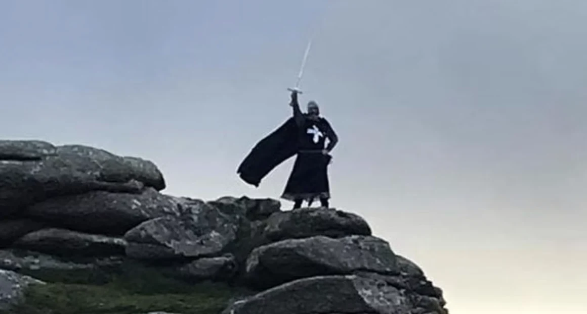 A knight in a black cloak raising a sword on granite rocks on the Cornish moor