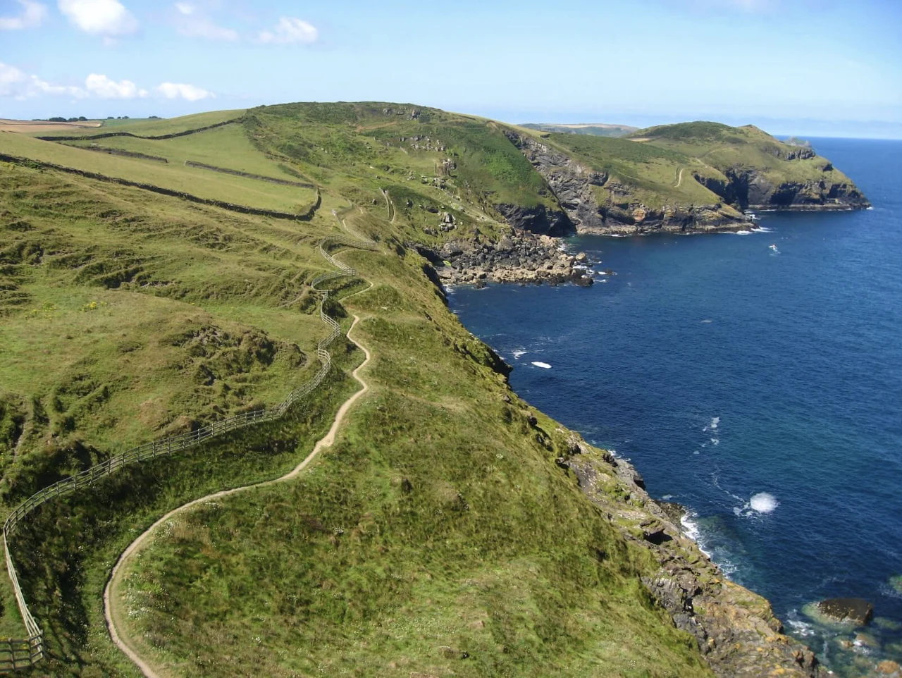 An aerial view of a fenced coast path winding along cliffs near Pentire Point, a rocky headland and blue sea beyond