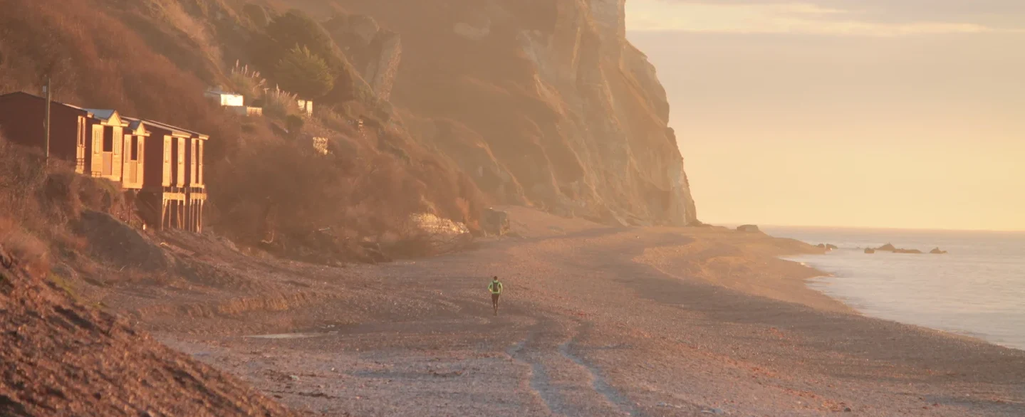 A jogger running along the shingle at Branscombe Beach in Devon at low sun, beach huts and chalk cliffs behind. Photo: National Trust / Chris Mason