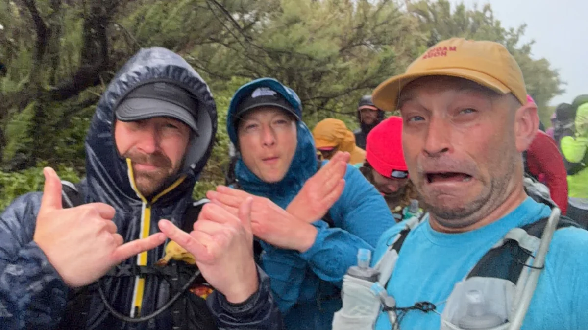 Three PZ×RC runners in hooded jackets on the coast path in heavy rain, pulling faces at the camera